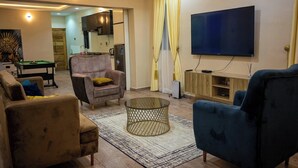 A living room with a pool table, a blue armchair, a sofa, a television, and a kitchen area in the background. - Nomsy Luxury Service Apartments  (Owerri)