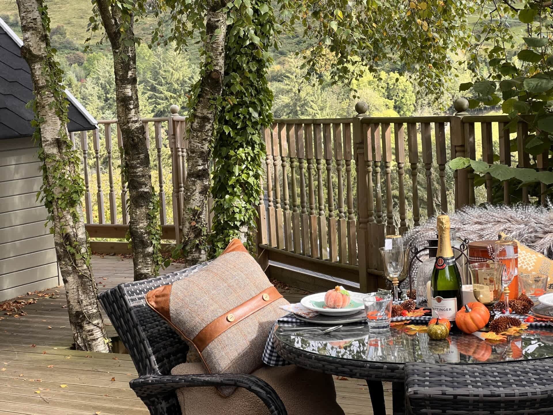 An outdoor dining area with a table set for two, surrounded by a wooden deck and a view of a forested landscape.