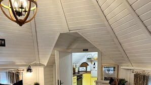 A cozy attic bedroom with a bed, a dining table, and a chair. There is a kitchen area visible through an open door.