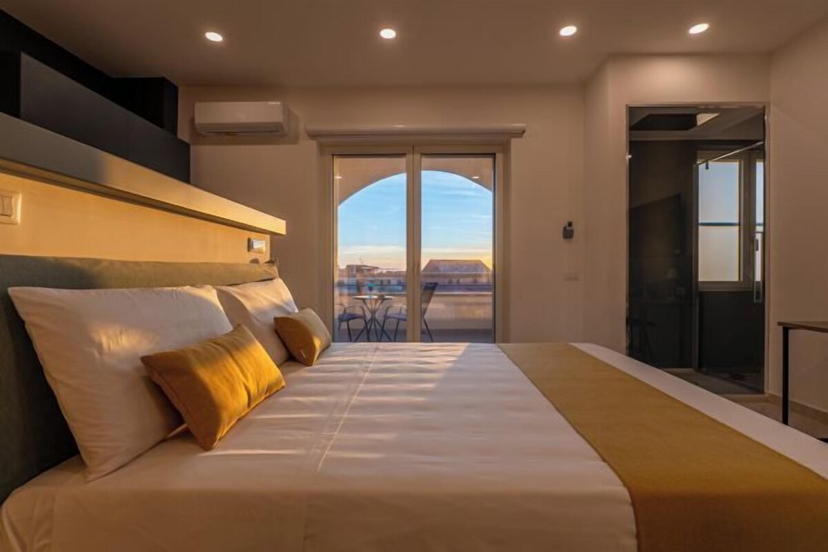 A hotel room with a large bed, a view of the city through a glass door, and a balcony with outdoor furniture.