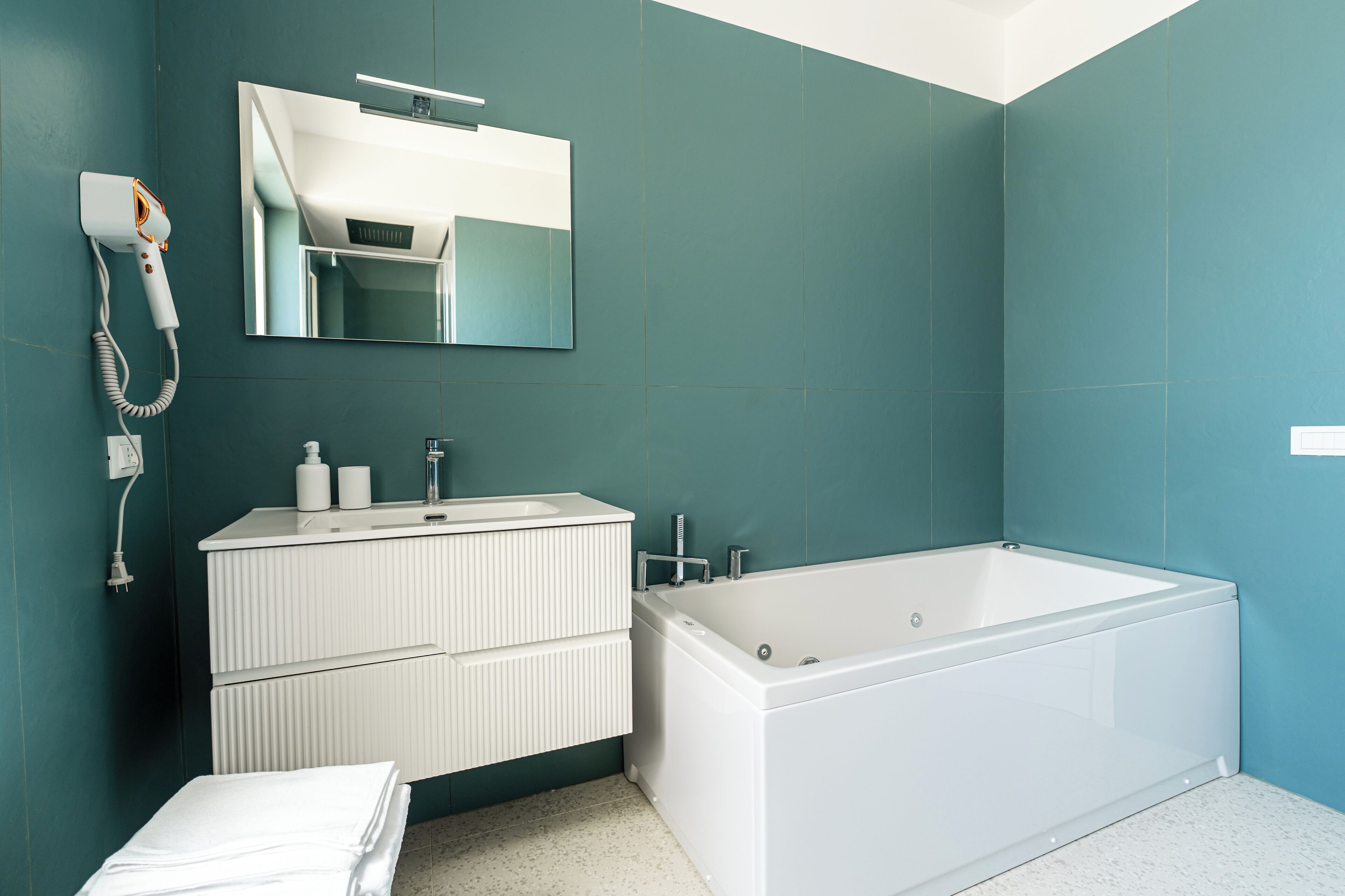 A modern bathroom with teal walls, a white vanity with a mirror, a white bathtub, and a towel on the floor.