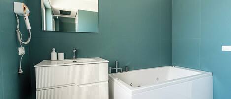 A modern bathroom with teal walls, a white vanity with a mirror, a white bathtub, and a towel on the floor.