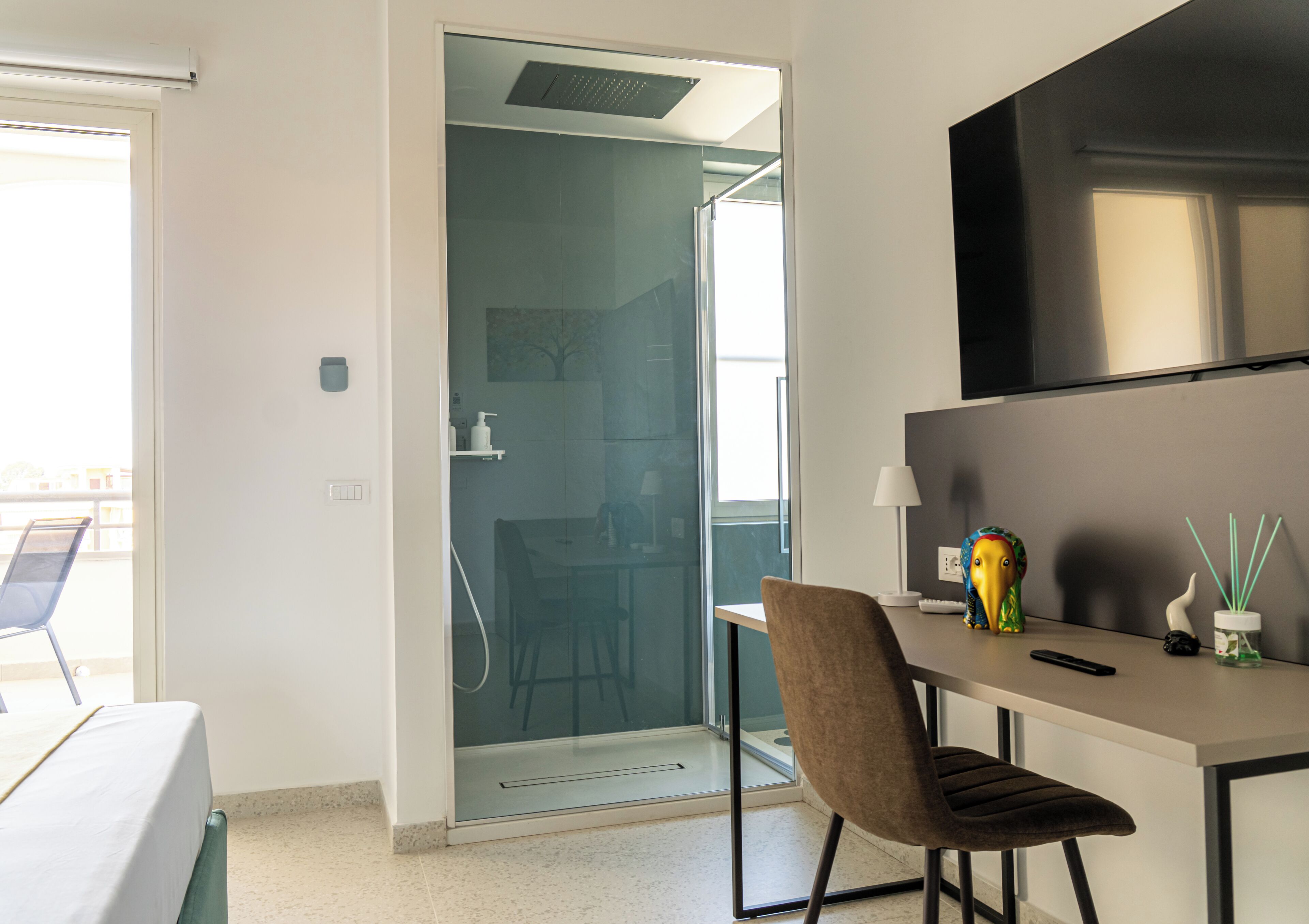 A modern hotel room with a glass door leading to a shower area, a desk with a chair, and a mounted TV.