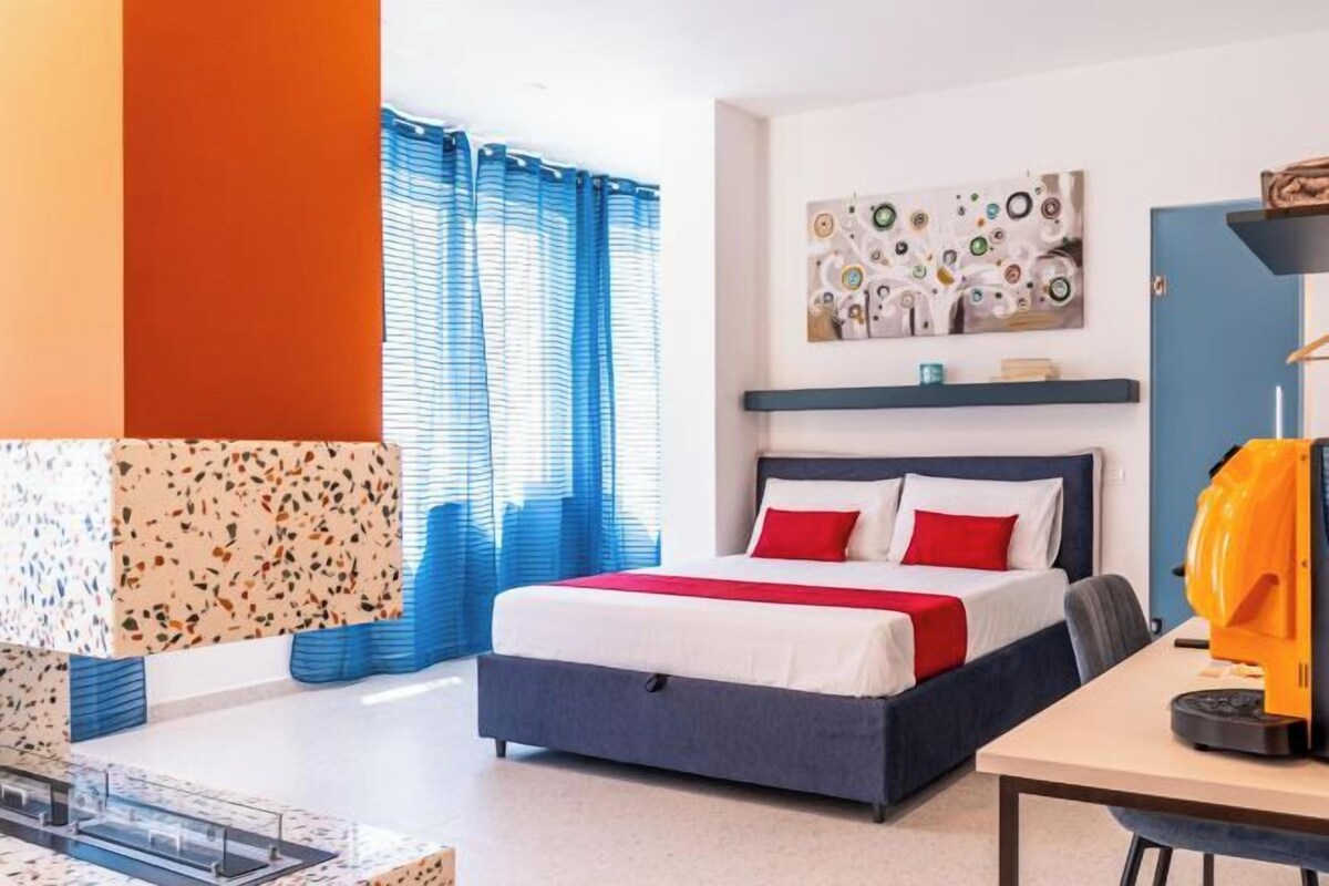 A hotel room with a bed, a desk, a chair, a window with blue curtains, and a wall with a textured design.