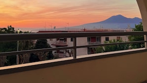 Un balcón con vista a una puesta de sol sobre una montaña, una mesa con un objeto negro y un objeto cilíndrico rojo.
