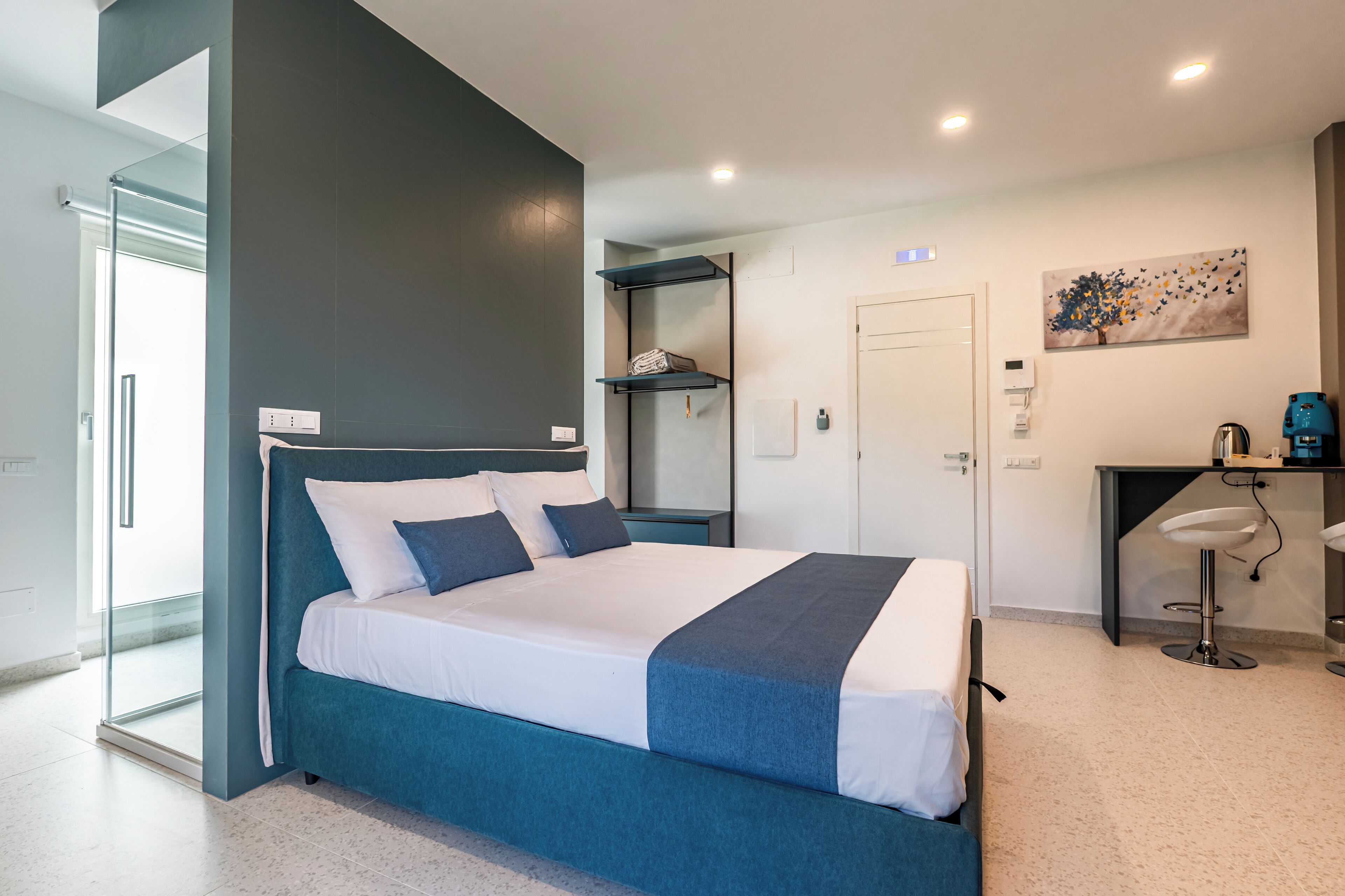 Une chambre moderne avec un lit, une porte vitrée donnant sur un balcon, un petit bureau avec une chaise et une étagère fixée au mur.