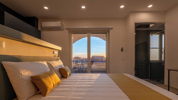 Habitación de hotel con una cama grande, vista a la ciudad a través de un ventanal y un balcón con mobiliario exterior.