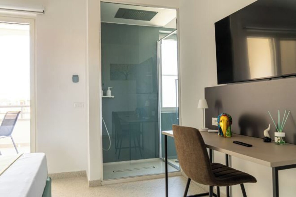 Habitación de hotel moderna con puerta de vidrio que da a un balcón, una pequeña mesa con una silla y un televisor montado en la pared.