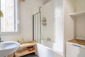Ein Badezimmer mit einer weißen Badewanne, einem hölzernen Ablagefach mit einem Kupferbecken, einem weißen Waschbecken und einem Fenster.