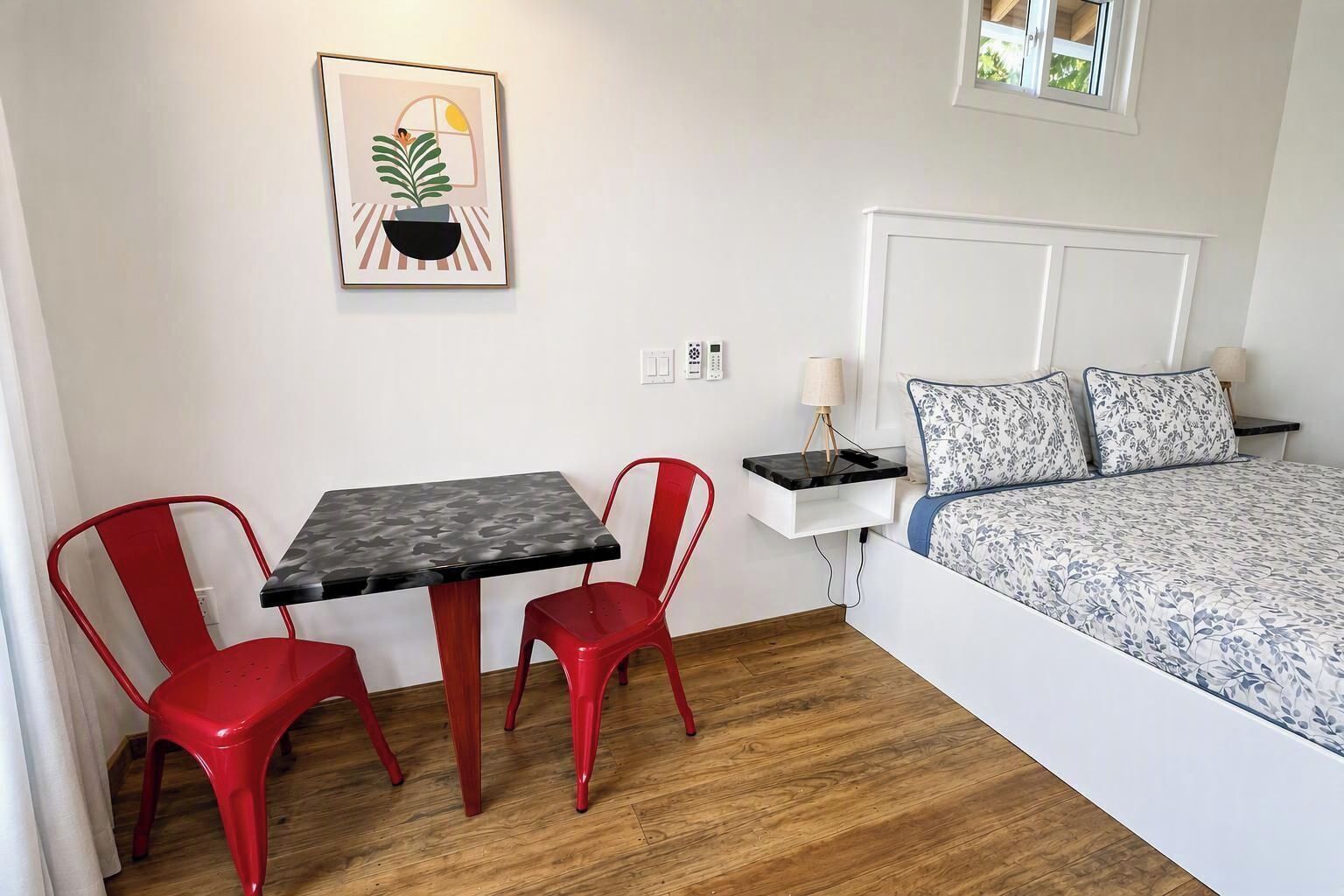 Una habitación pequeña y limpia con una cama, una mesita de noche, una lámpara, un cuadro enmarcado y dos sillas rojas alrededor de una mesa.