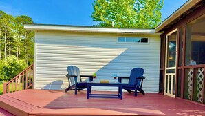A covered deck with two Adirondack chairs and a small table, overlooking a white building with a window and a door.