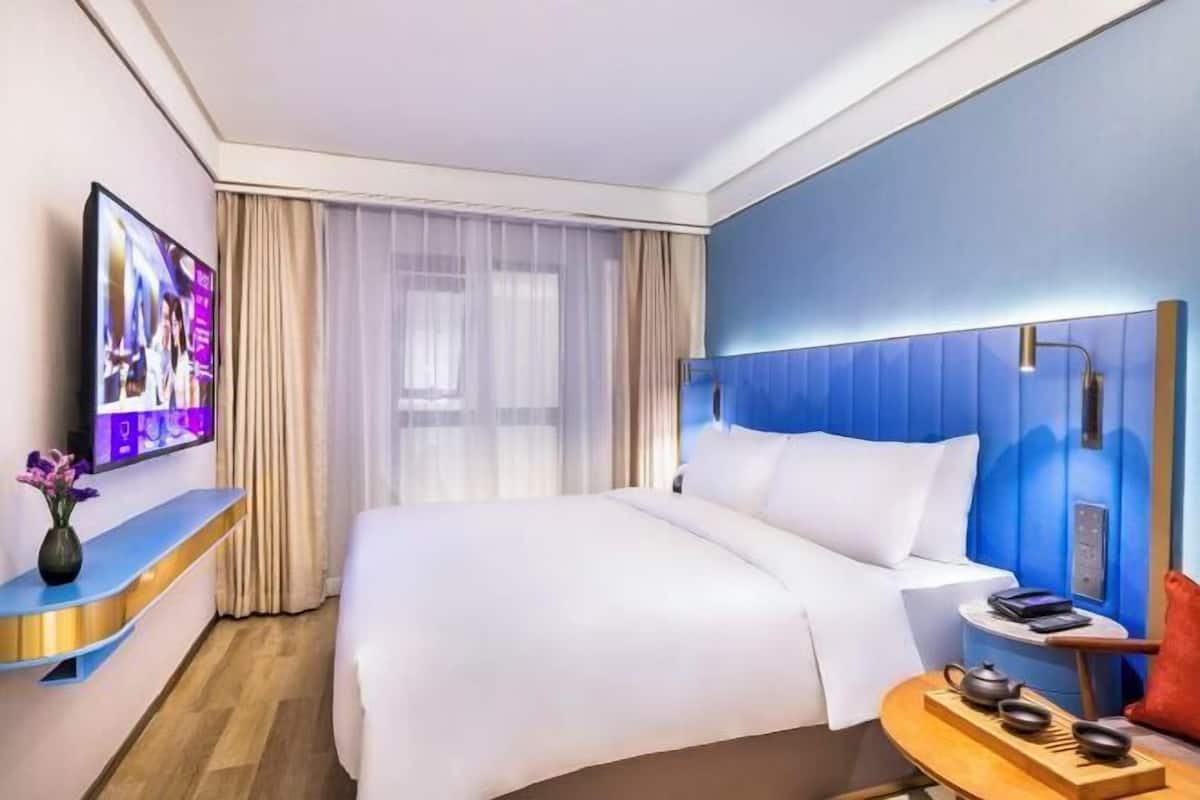 Una habitación de hotel moderna con una cama grande, un televisor en un soporte, una mesita de noche con una lámpara y vistas a la ciudad a través de la ventana.
