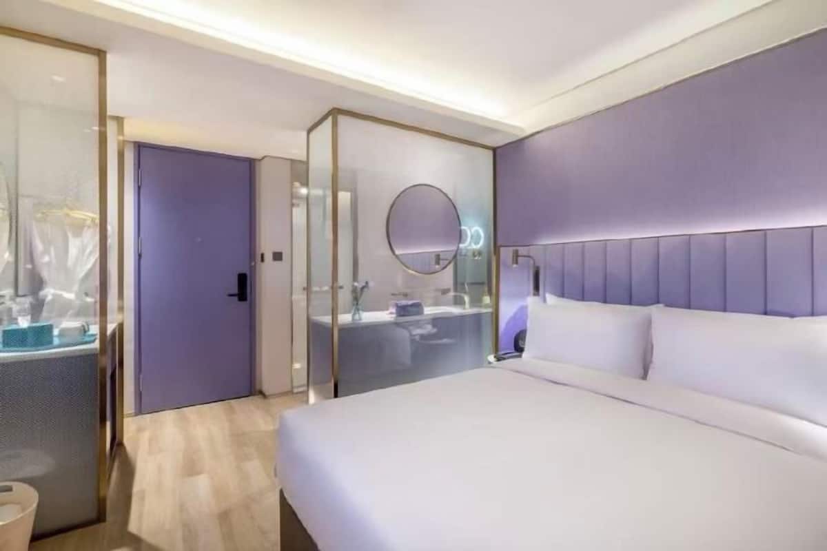 Una habitación de hotel moderna con una cama grande, un separador de vidrio, un baño con espejo y una lámpara fijada a la pared.