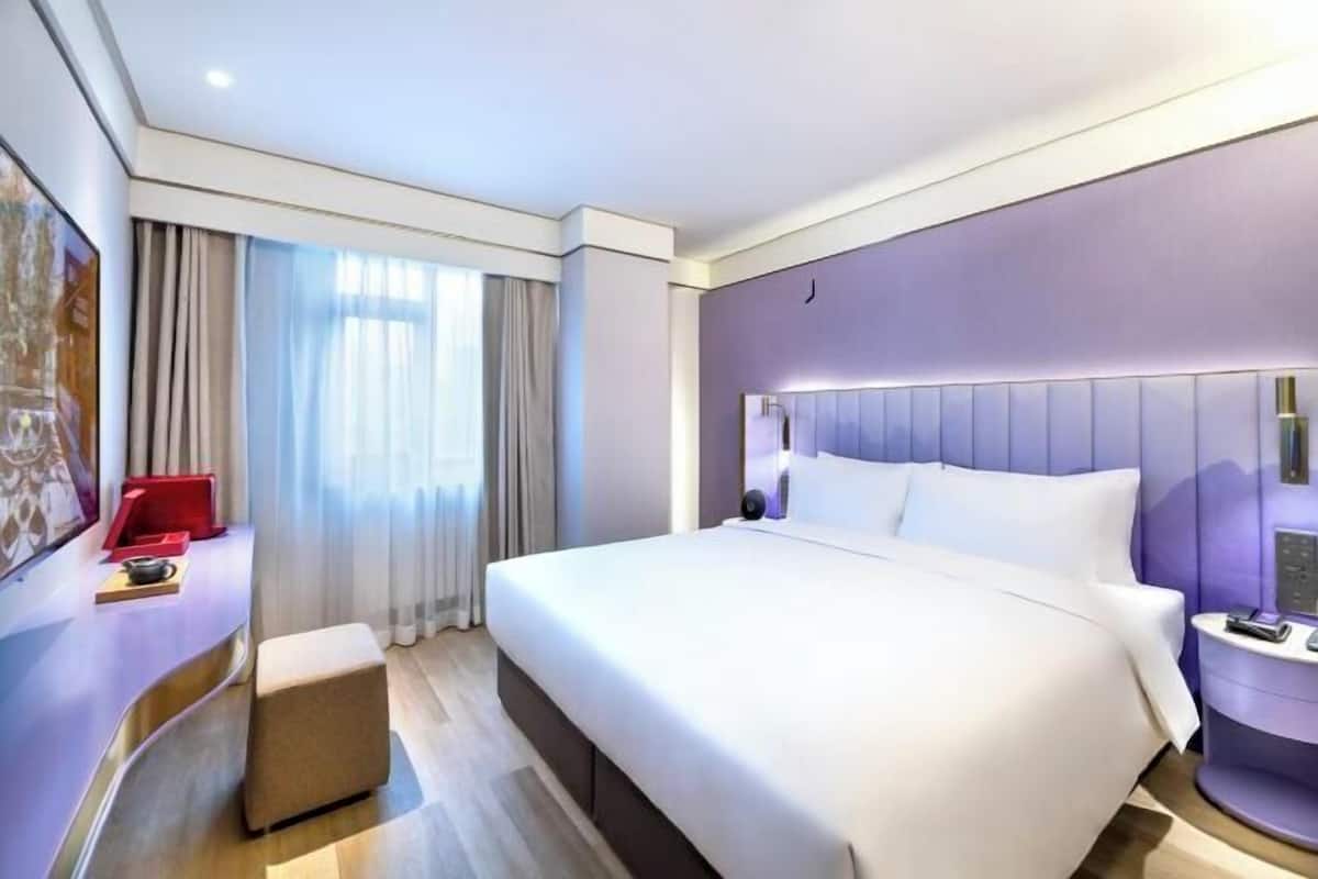 Una habitación de hotel moderna con una cama grande, una mesita de noche, una silla roja y una ventana con cortinas.