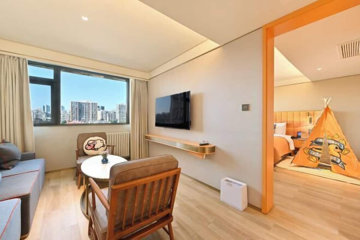 Una habitación de hotel con vistas a la ciudad, una mesa redonda con sillas, un televisor montado en la pared y una estructura similar a una tienda de campaña con un personaje de dibujos animados.