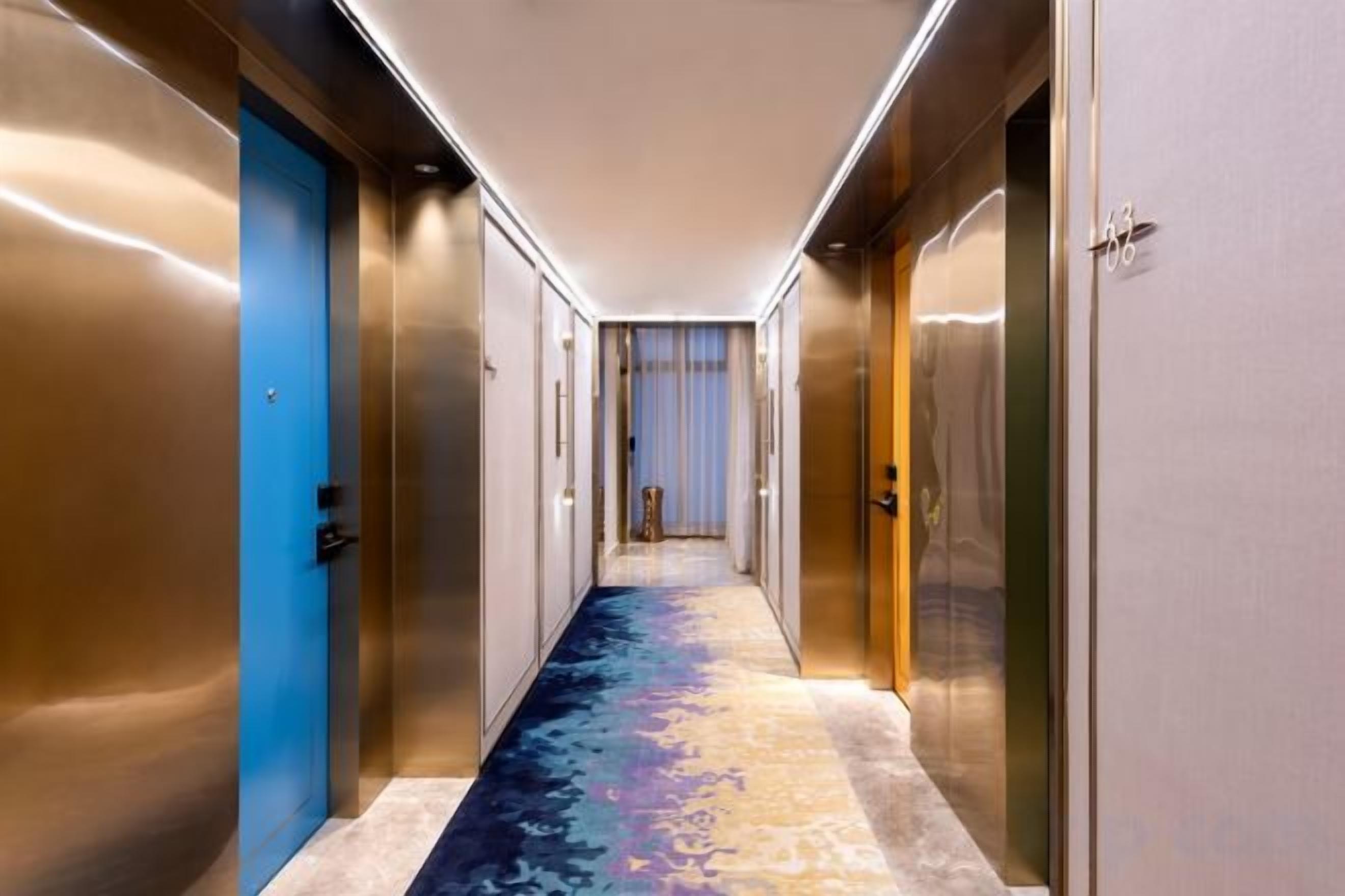 A hotel corridor with metallic doors, a carpeted floor, and a view into a room with a bed and curtains.