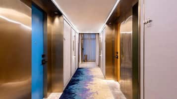 A hotel corridor with metallic doors, a carpeted floor, and a view into a room with a bed and curtains.