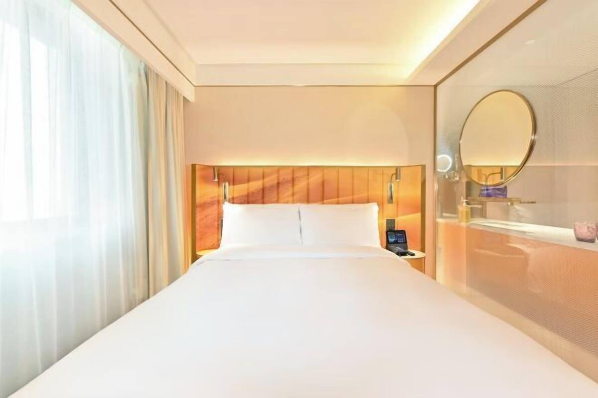 Habitación de hotel con una cama grande, una mesita de noche con teléfono, un espejo redondo y una ventana con cortinas.