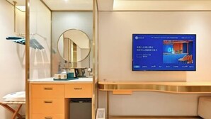 A modern hotel room with a wooden desk, a mirror, a television mounted on the wall, and a small cabinet.
