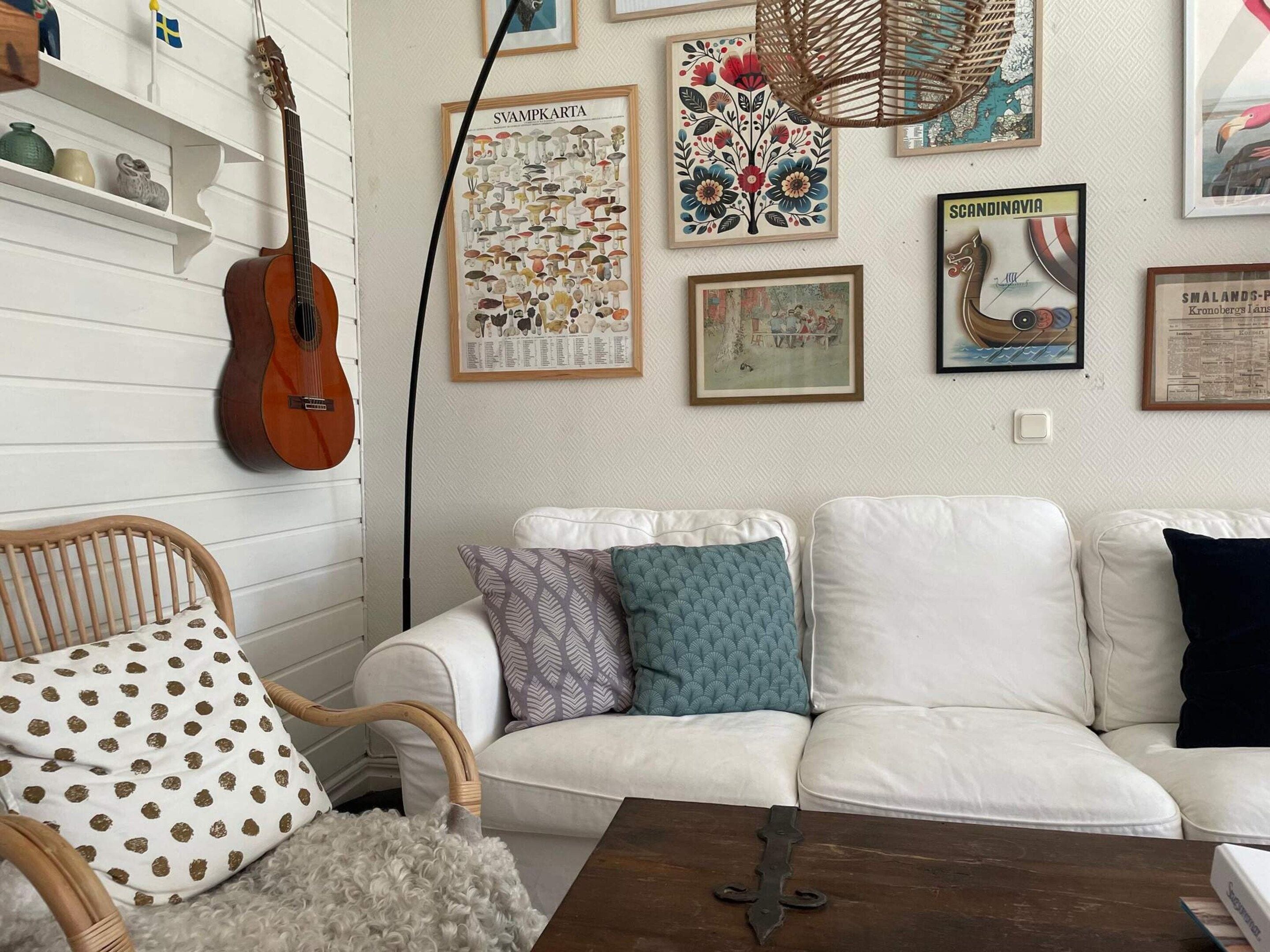 A living room with a white sofa, patterned cushions, a wicker chair, a guitar, and various framed pictures on the wall.
