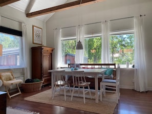 Ein Essbereich mit Tisch und Stühlen, einem Holzschrank, einem Korbwägelchen und einem großen Fenster mit Vorhängen.