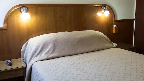 Een hotelkamer met een houten hoofdbord, twee bedlampjes, een bed met een beddegoed met een patroon, en een nachtkastje met een afstandsbediening.