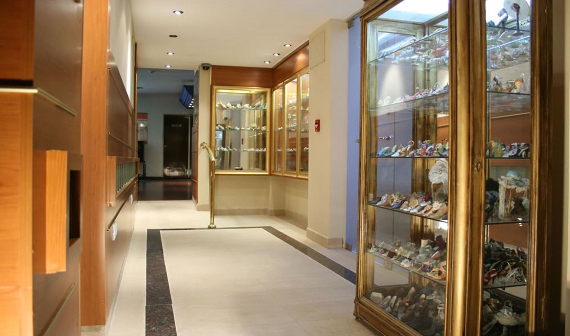 A hotel corridor with wooden paneling, a glass display case filled with shoes, and a reception area in the distance.