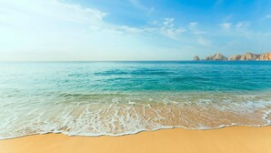 A beach with clear blue water, sandy shore, and rocky formations in the distance. - Serene 1BR Beachfront Stay with Outdoor Pool (Cabo San Lucas)