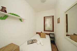 A small, clean room with a bed, a shelf with decorative items, a window with curtains, and a mirror. - La casa della Bougan-ville (Toscolano-Maderno)