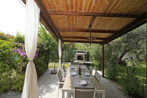 An outdoor dining area with a wooden pergola, a table set for four, and a pizza oven in the background. - La casa della Bougan-ville (Toscolano-Maderno)