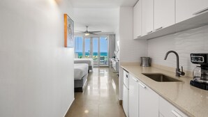 A modern hotel room with a kitchenette, a bed, a ceiling fan, and a view of the ocean.