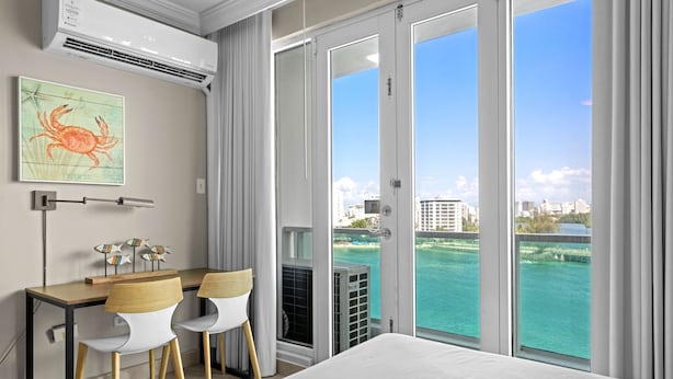 Habitación de hotel con cama, escritorio con dos sillas, aire acondicionado, ventana con vistas al mar y edificios, y un cuadro en la pared.