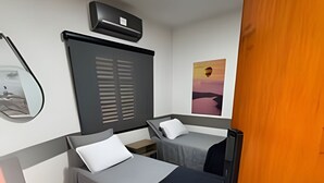 Une petite chambre à coucher, de taille réduite, avec deux lits simples, une table de chevet, un miroir, un climatiseur fixé au mur et un tableau encadré accroché au mur.