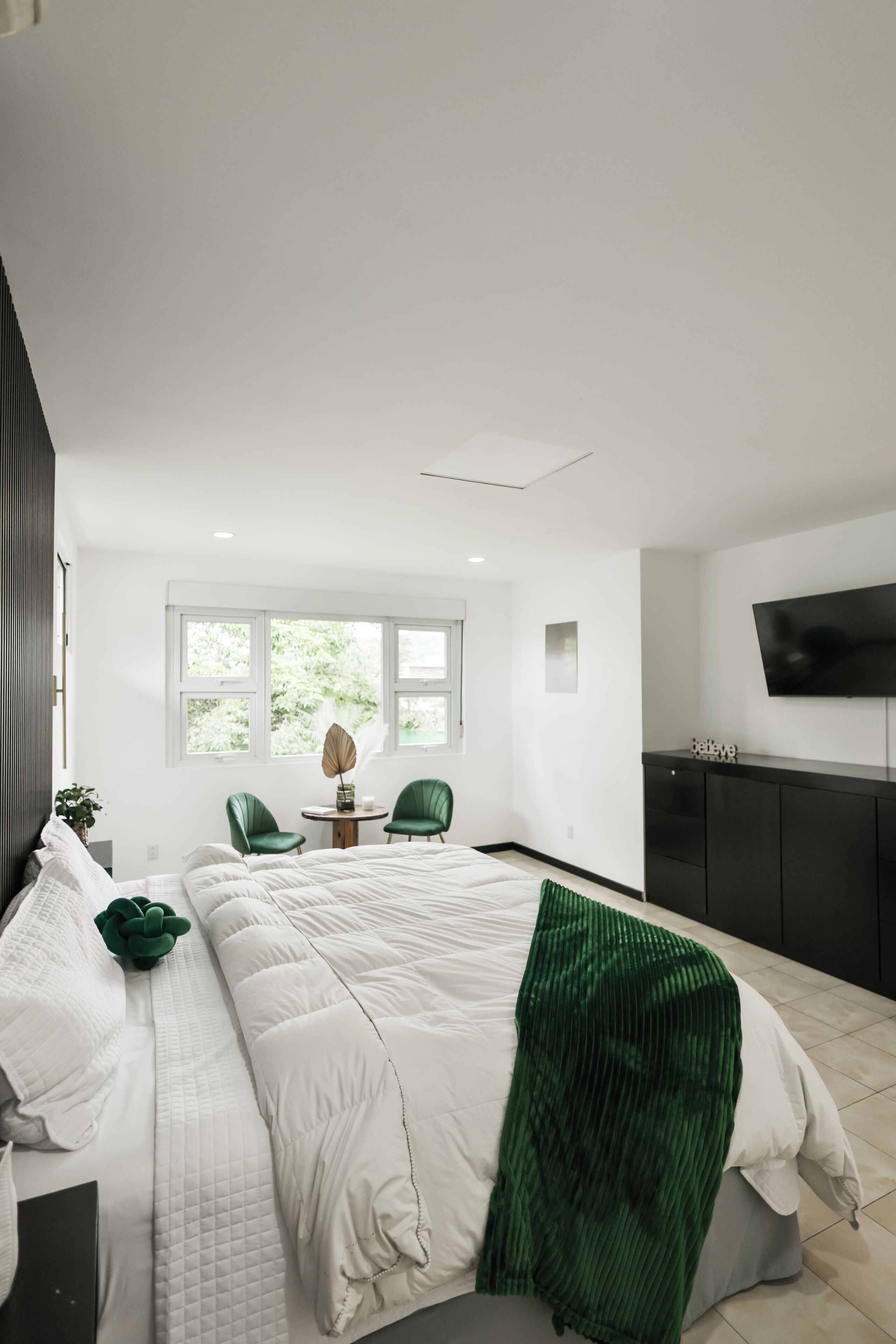 A modern bedroom with a large bed, green chairs, a small table, and a TV on the wall.