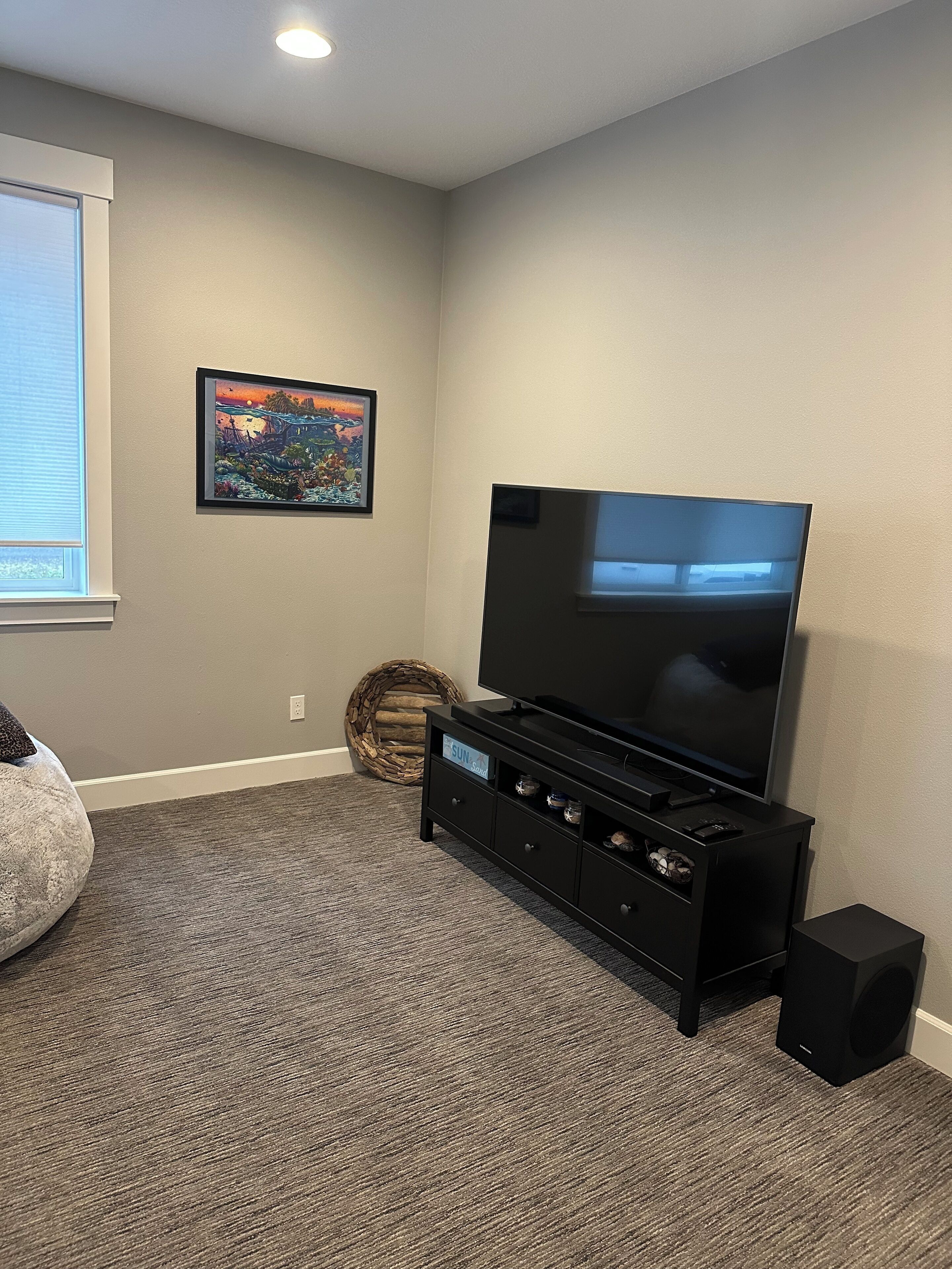 Ein Wohnzimmer mit einem Flachbildfernseher auf einem schwarzen Standfuß, einem grauen Teppich, einem gerahmten Bild an der Wand und einem Korbwäschesack.