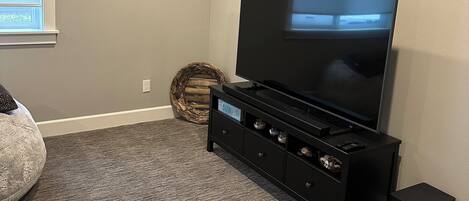 Ein Wohnzimmer mit einem Flachbildfernseher auf einem schwarzen Standfuß, einem grauen Teppich, einem gerahmten Bild an der Wand und einem Korbwäschesack.