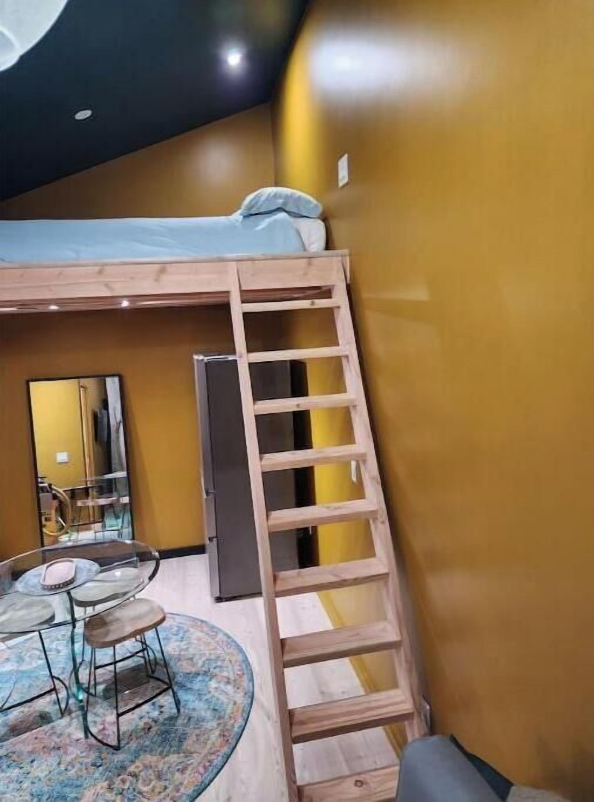 Ein kleines Zimmer mit Etagenbett, einer Leiter, einem kleinen Tisch mit Stühlen und einem Kühlschrank.