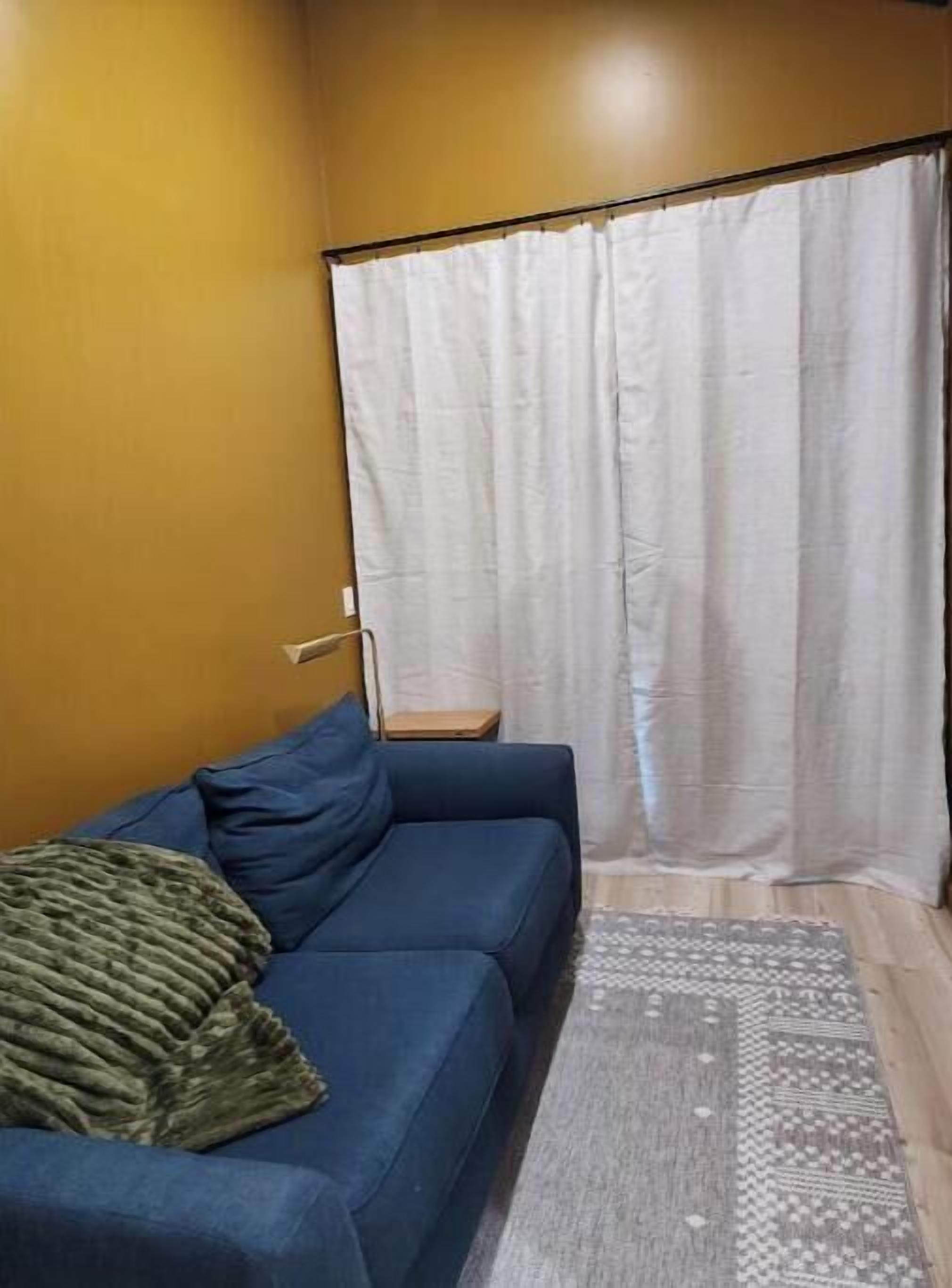 Una stanza con un divano blu, un piccolo tavolino di legno, un tappeto a fantasia e una tenda bianca.