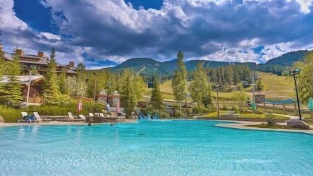Ein klares, blaues Schwimmbecken mit Liegestühlen, umgeben von Bäumen und Bergen im Hintergrund.