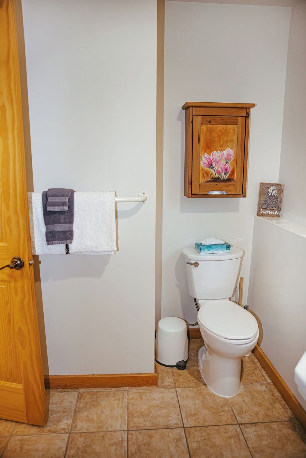 Une salle de bain comprenant des toilettes, une poubelle, un porte-serviettes avec une serviette, un meuble en bois orné d’un tableau floral et une pancarte indiquant « chez soi ».
