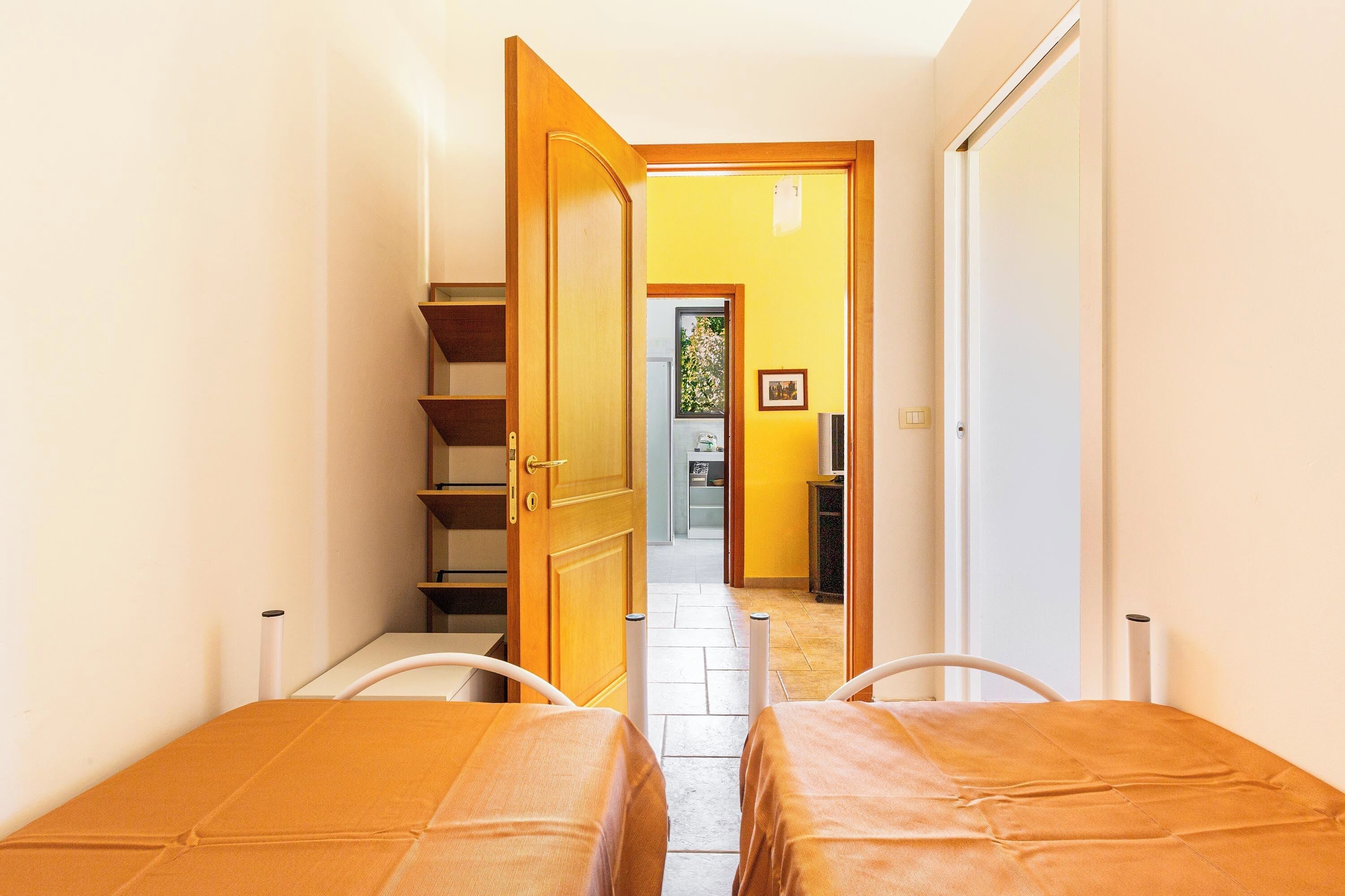 Een smalle gang met twee bedden, een houten deur en zicht op een andere kamer met een raam.