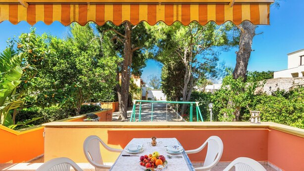 An outdoor patio with a table set for dining, surrounded by chairs, and a railing overlooking a garden.