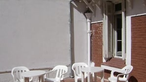 Une terrasse avec des chaises en plastique blanches et une petite table, située à côté d’un bâtiment dont le mur est en briques et la porte est blanche.