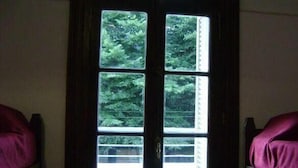 Une pièce dotée d’une grande fenêtre divisée en quatre panneaux, chacun offrant une vue sur un feuillage dense. On y trouve deux lits avec une literie rose.