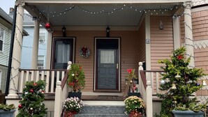 Ein zweistöckiges Haus mit Veranda, verziert mit Weihnachtsbeleuchtung und Dekorationen, einem kleinen Weihnachtsbaum und Blumentöpfen.