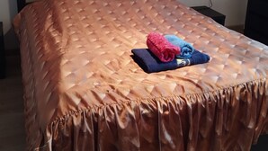 A bed with a brown quilted cover and a ruffled edge, a folded pink and blue towel, and a wall-mounted light fixture.