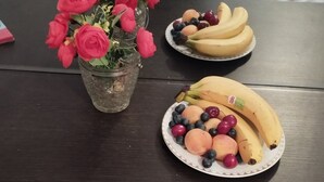 A table with a vase of artificial flowers, two plates of fruit, and a bed with a brown cover in the background.