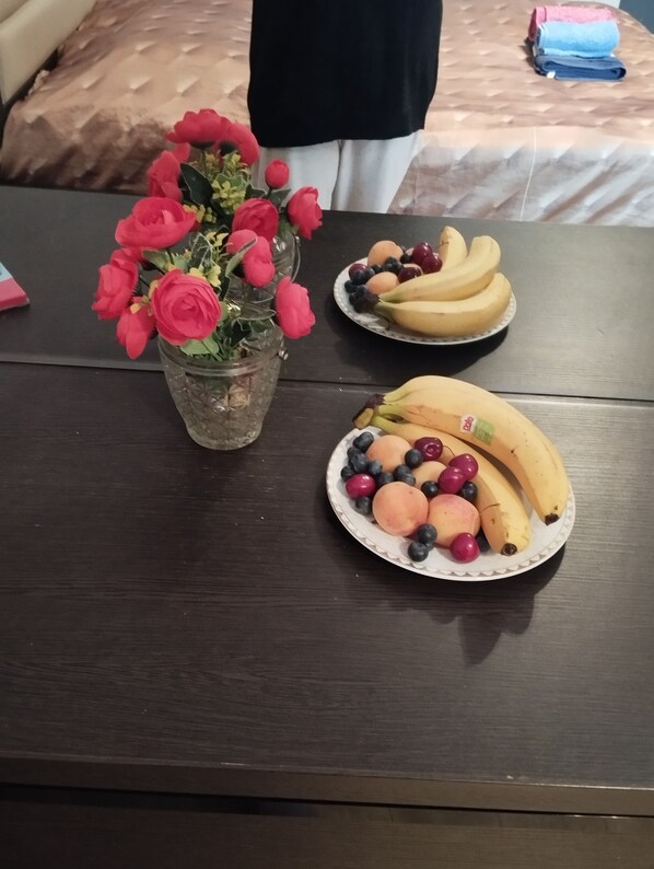 A table with a vase of artificial flowers, two plates of fruit, and a bed with a brown cover in the background.