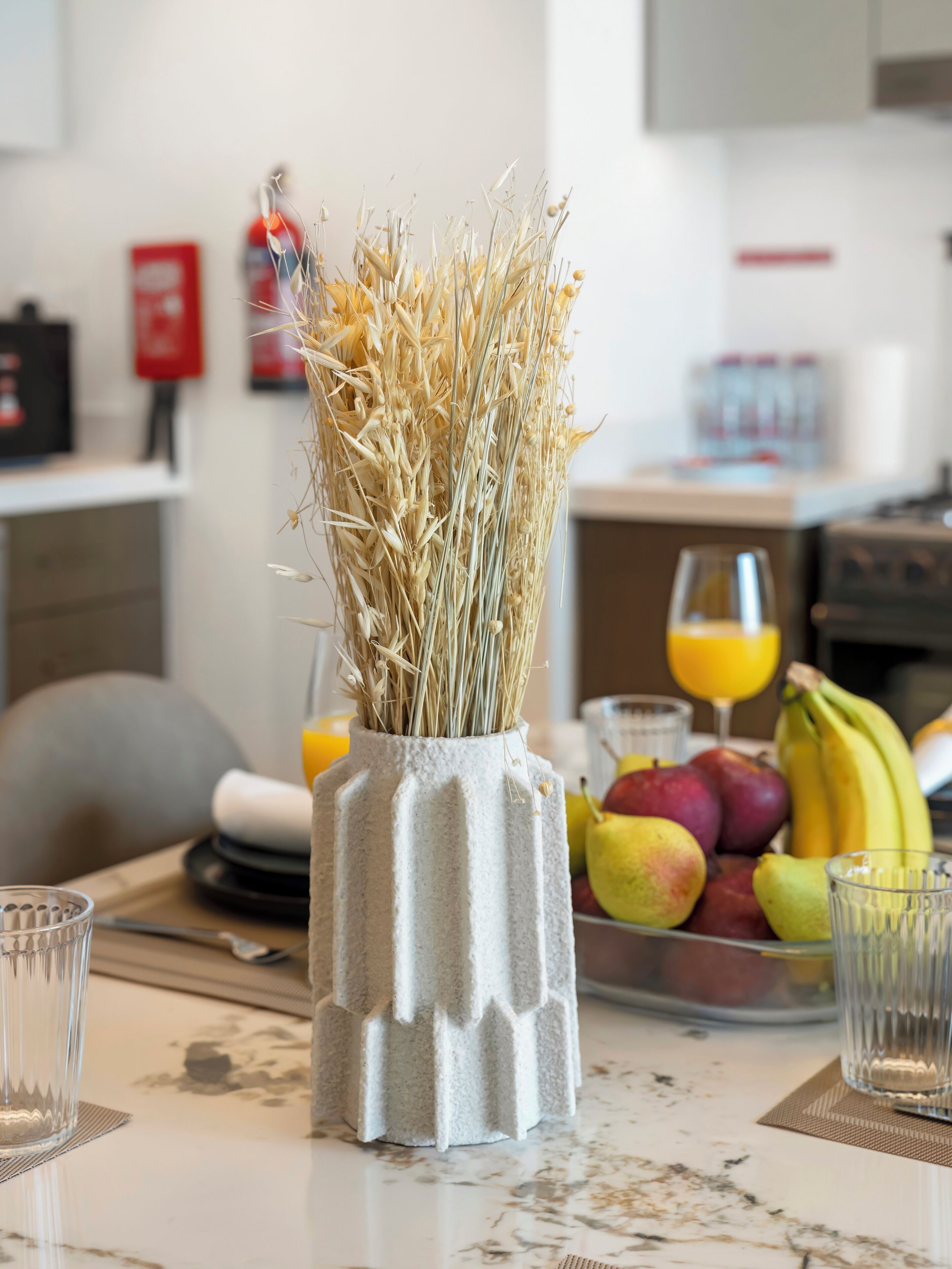 A kitchen counter with a vase of dried plants, a fruit bowl, and a glass of orange juice.