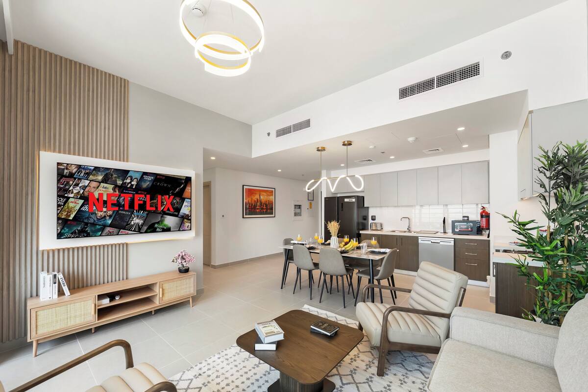 Un salon moderne doté d’un téléviseur à écran plat affichant le logo « NETFLIX », un coin repas avec une table dressée pour quatre personnes et une cuisine équipée d’appareils électroménagers modernes.